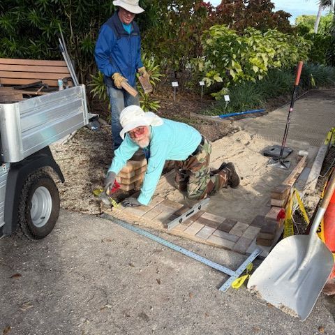 Two people laying bricks on a pathway outdoors. One kneels and works with tools; the other holds a hammer.