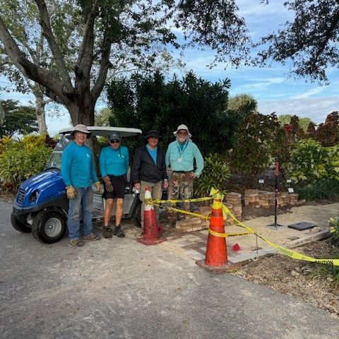 Four people stand near a blue golf cart, construction cones, and caution tape. They are in a garden setting.