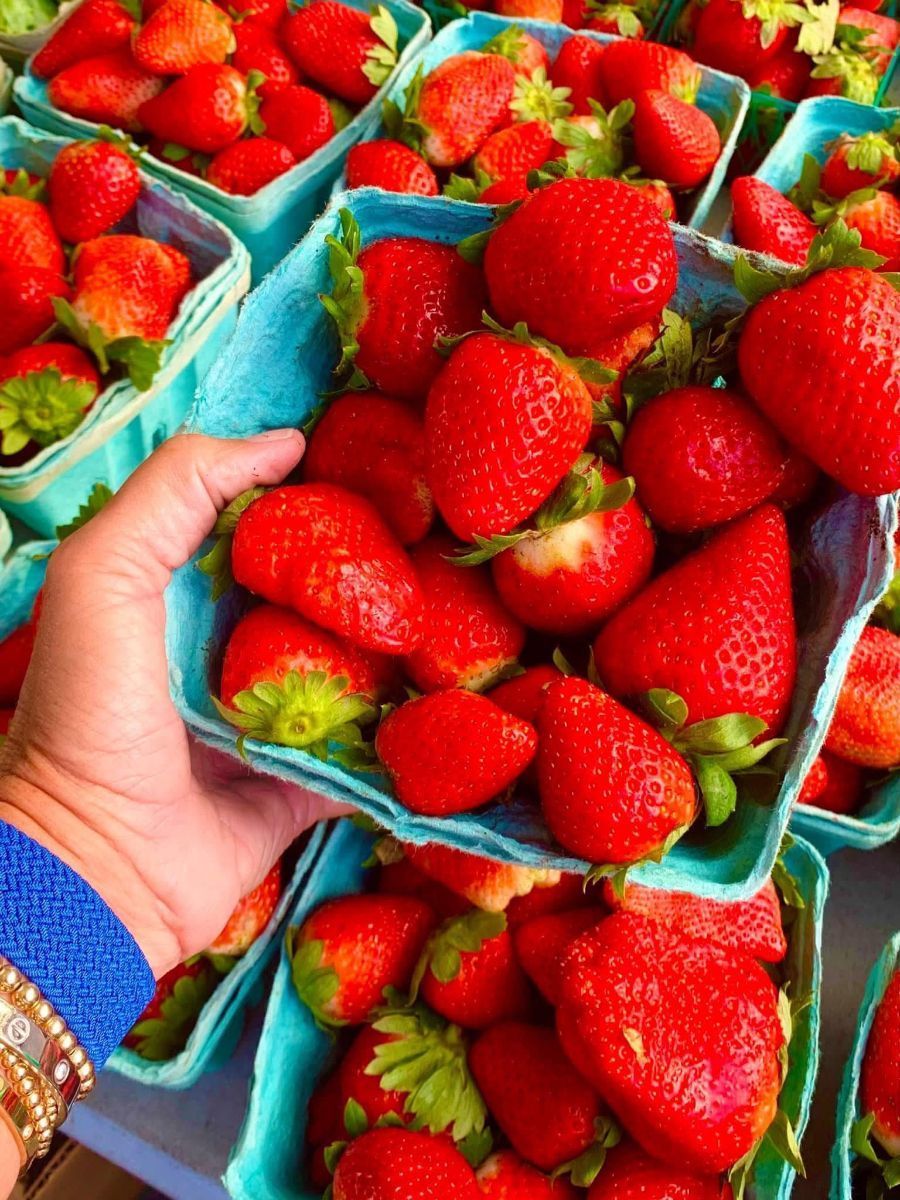 Hand holding a full container of ripe red strawberries at a market. Other full containers are visible.