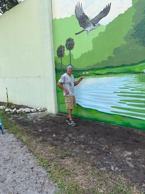 Man stands near a mural of an osprey over a lake, pointing. The wall is green and light yellow.