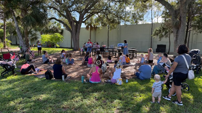 People gathered outdoors for an event under trees, some seated. Benches and a fence are in the background.