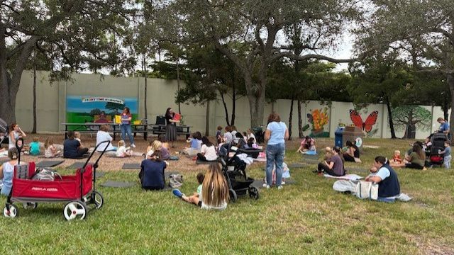 People sitting on grass at an outdoor event near a wall with murals. Red wagon in foreground.