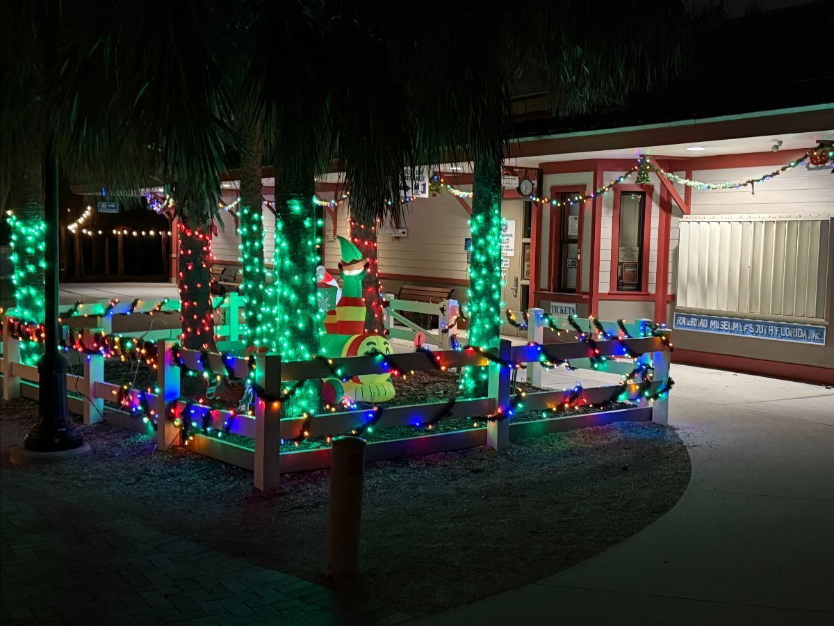 Christmas lights decorate a building's exterior with a fence, and palm trees. An inflatable elf is visible.