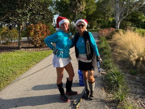 Two women in Santa hats and blue tops stand on a pathway, one in a skirt and boots, the other holding a leash.