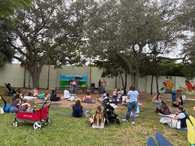 Group of people gathered outdoors for an event; presenter speaks in front of children and adults.