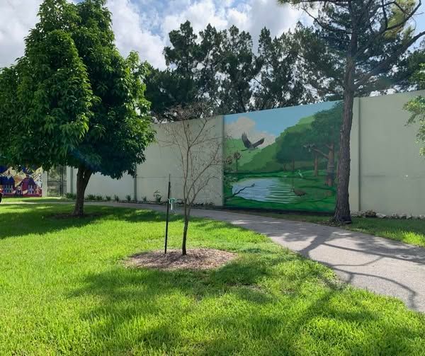 Green grassy park with a painted mural of a pond, birds, and trees on a white fence.