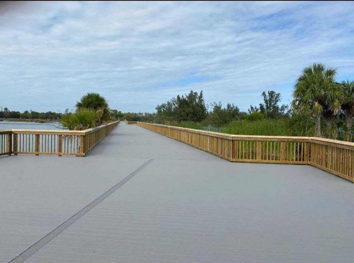 Boardwalk path with wooden railing, surrounded by greenery under a cloudy sky.