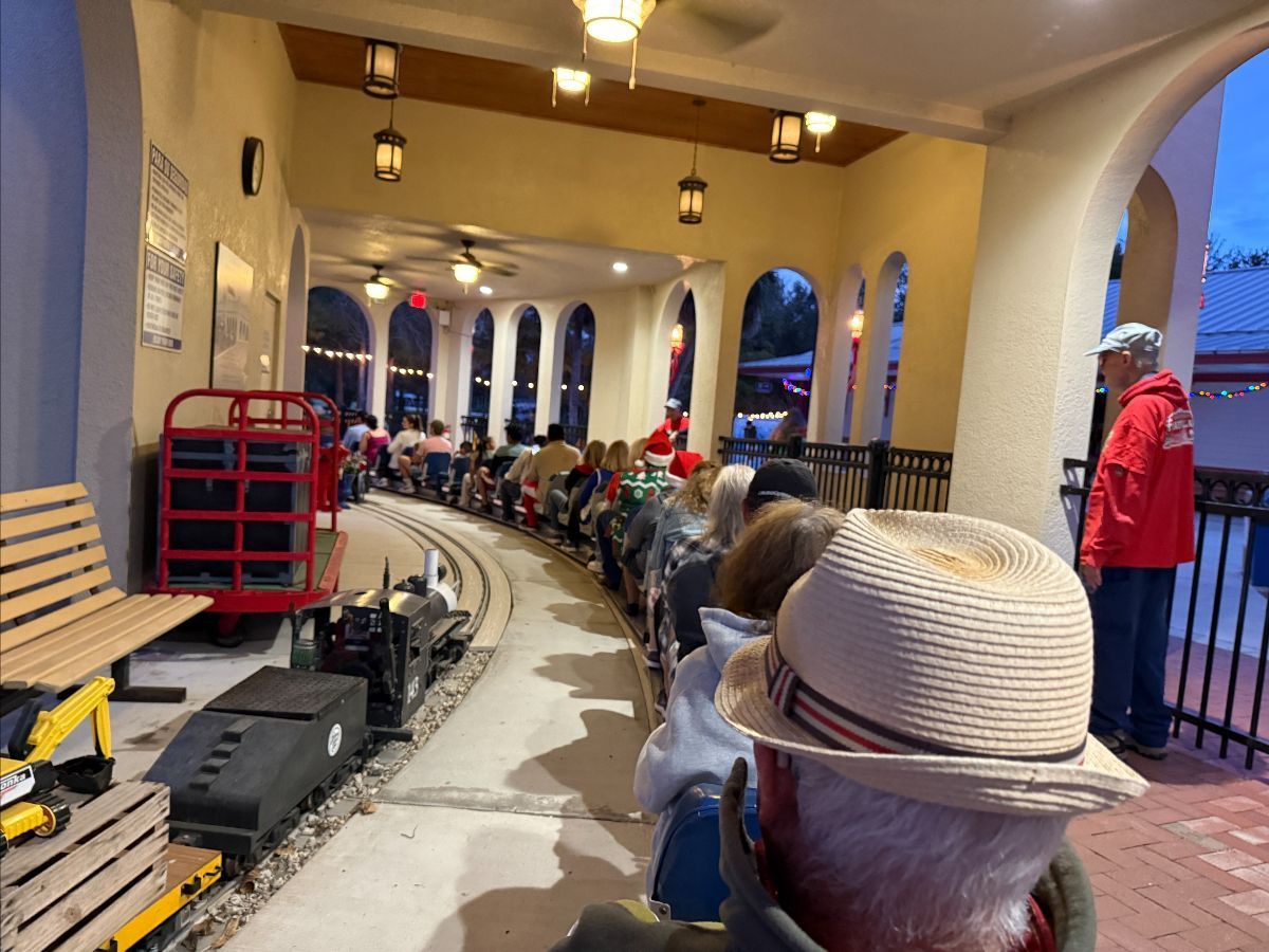 People ride a miniature train at an outdoor station with arches, a curved track, and hanging lights.
