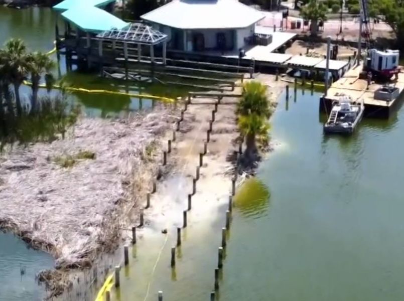 Aerial view of a dock construction site with a building, boats, and a sandy path by the water.