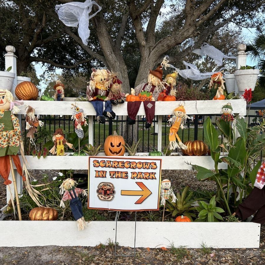 Halloween display: scarecrows on a white fence, pumpkins, sign