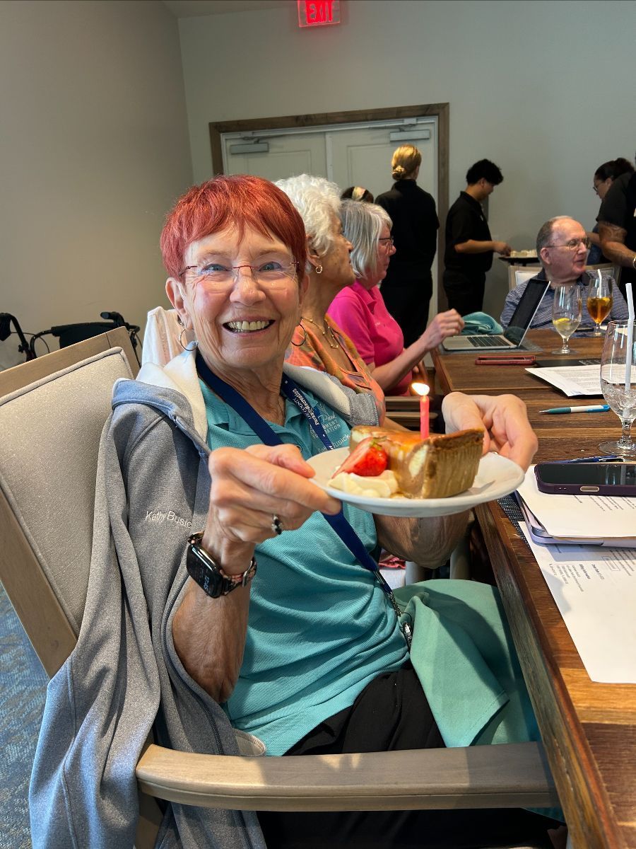 Woman holding a cake slice with a lit candle, smiling. Dining room setting, other people present.