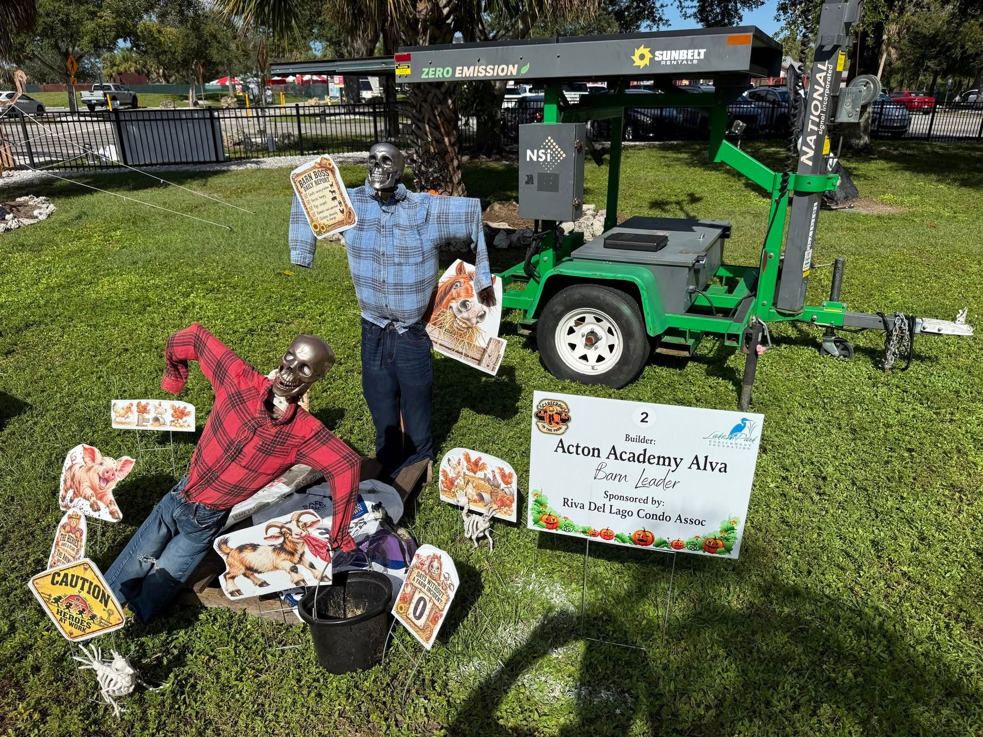Two scarecrow figures in a grassy outdoor display beside a green lawn mower and fall-themed signs