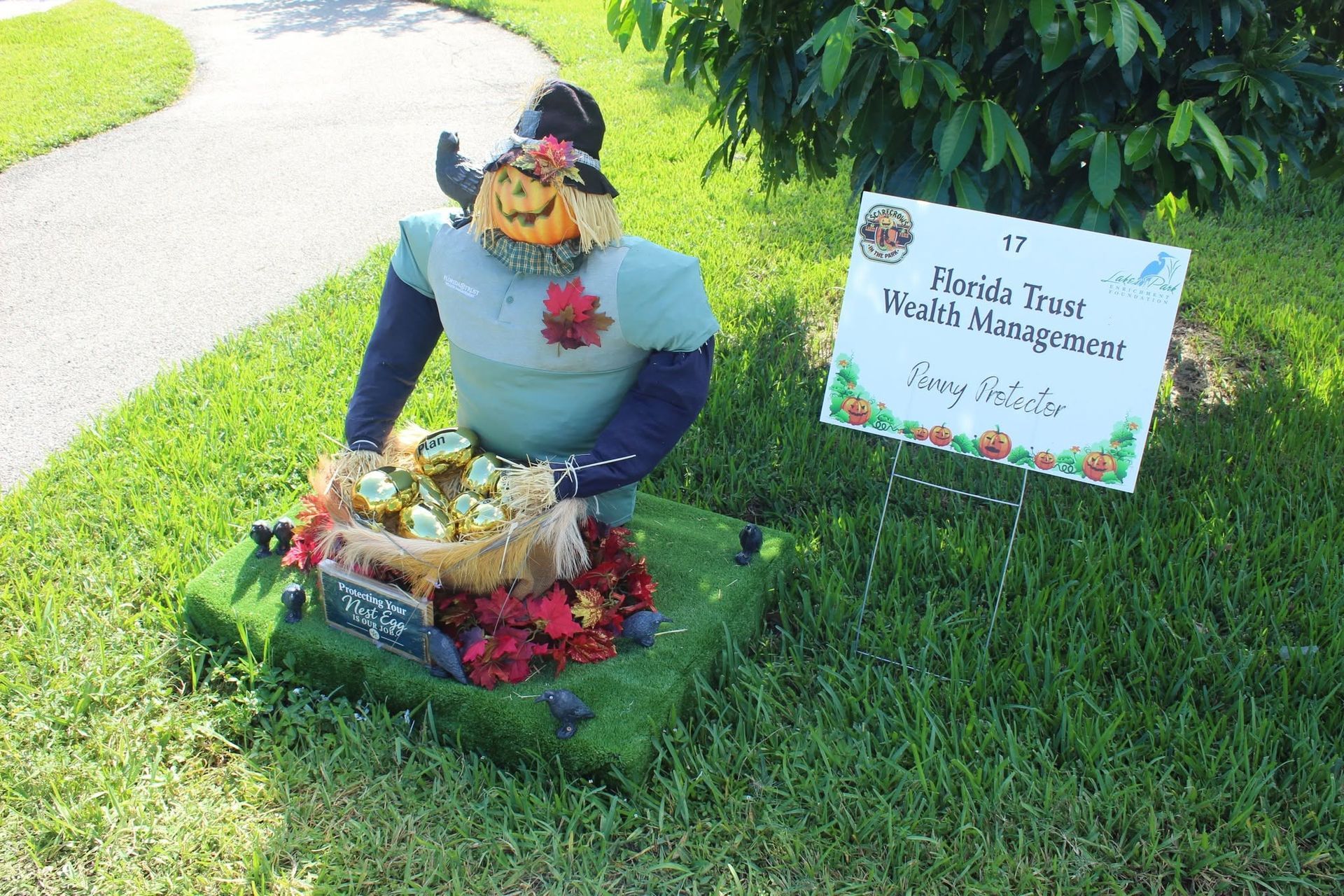 Scarecrow decoration beside a handwritten sign on a grassy lawn.