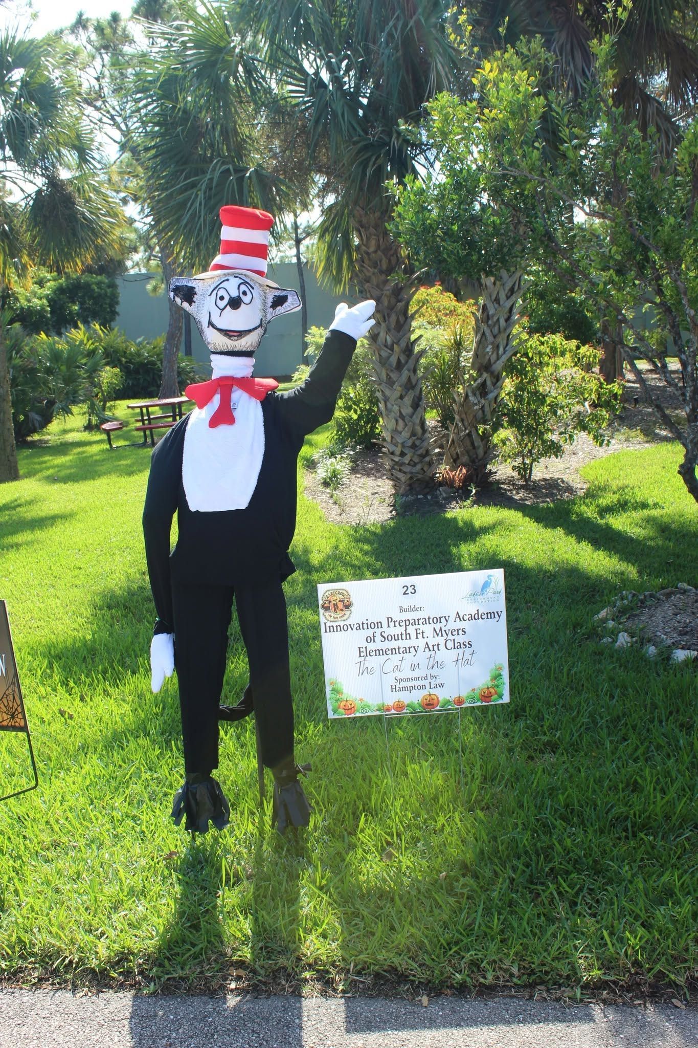 A cat mascot decoration in a red-and-white hat and bow tie stands on a grassy lawn by a sign.