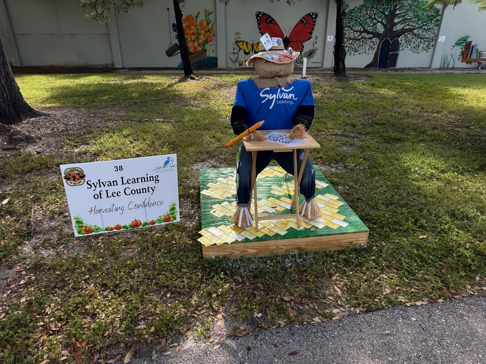Outdoor memorial display with a seated blue-shirted figure, butterfly wings, and a sign in a grassy yard.