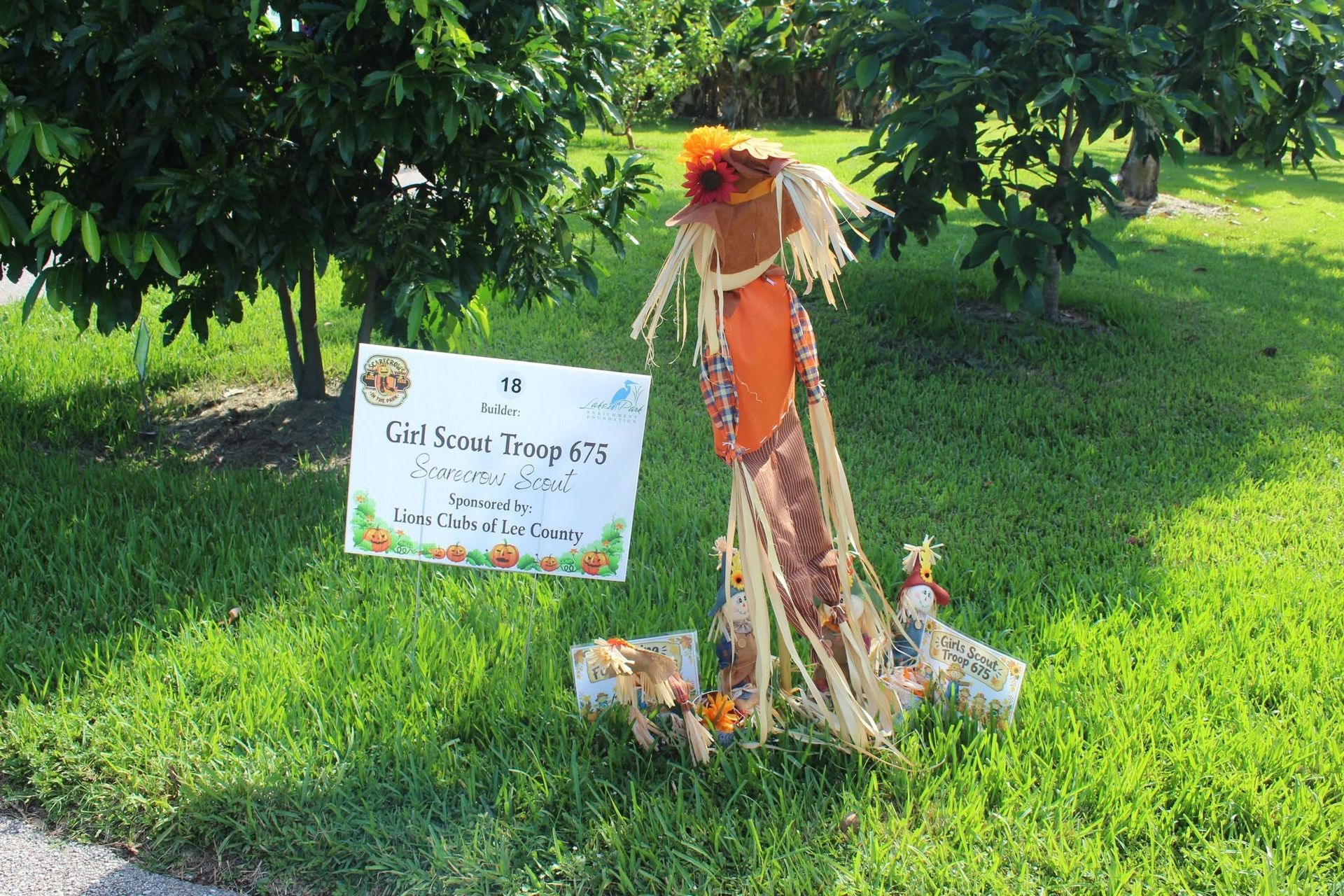 Scarecrow beside a sign in a sunny grassy yard under trees