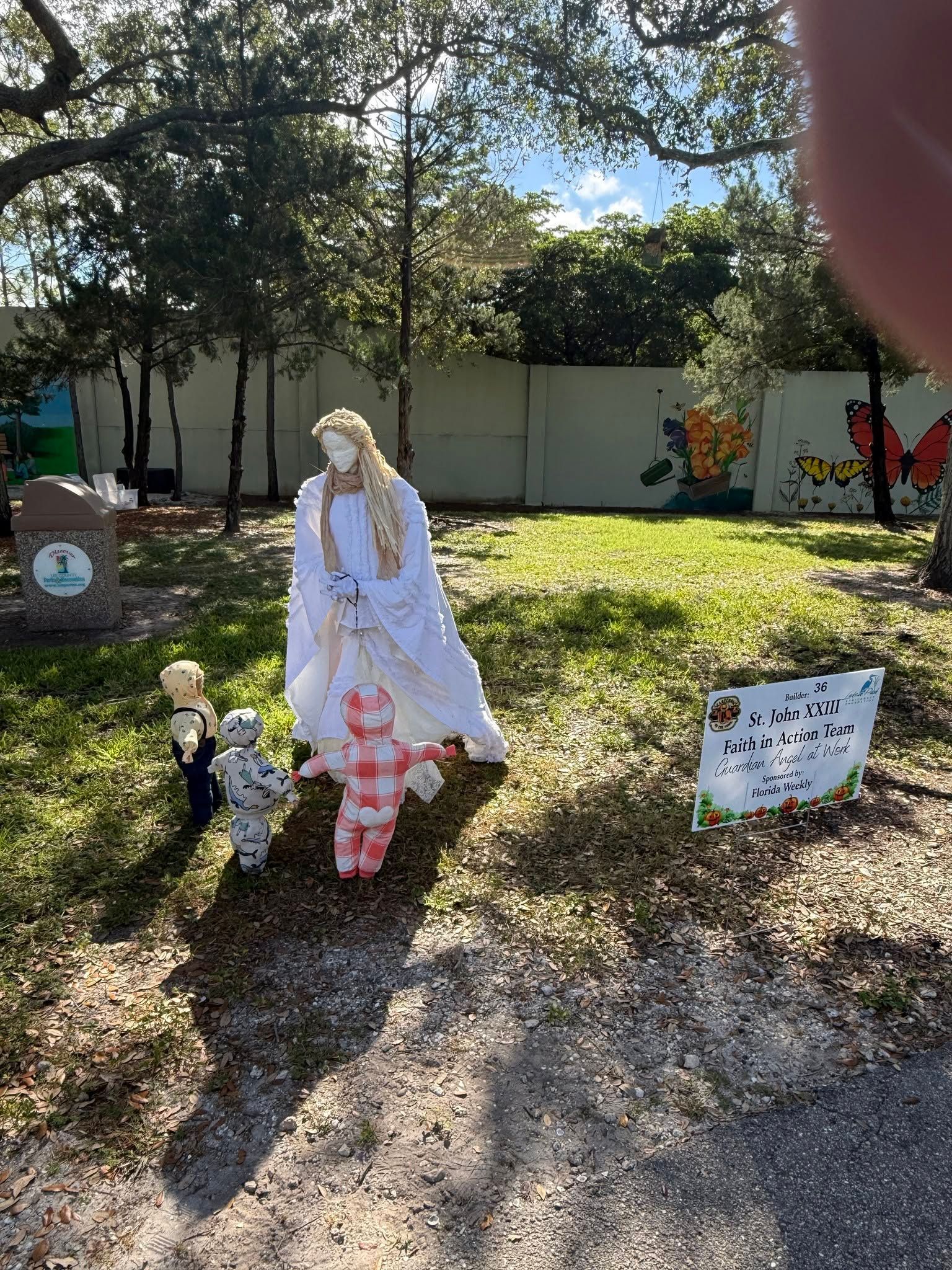 Outdoor garden display with a white-clad figure, small dog figures, and yard signs in sunlight