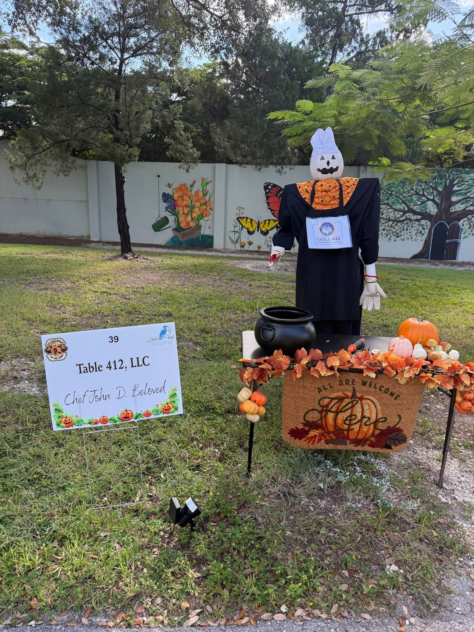 Halloween lawn display with a scarecrow, pumpkins, a sign, and autumn decorations outdoors.