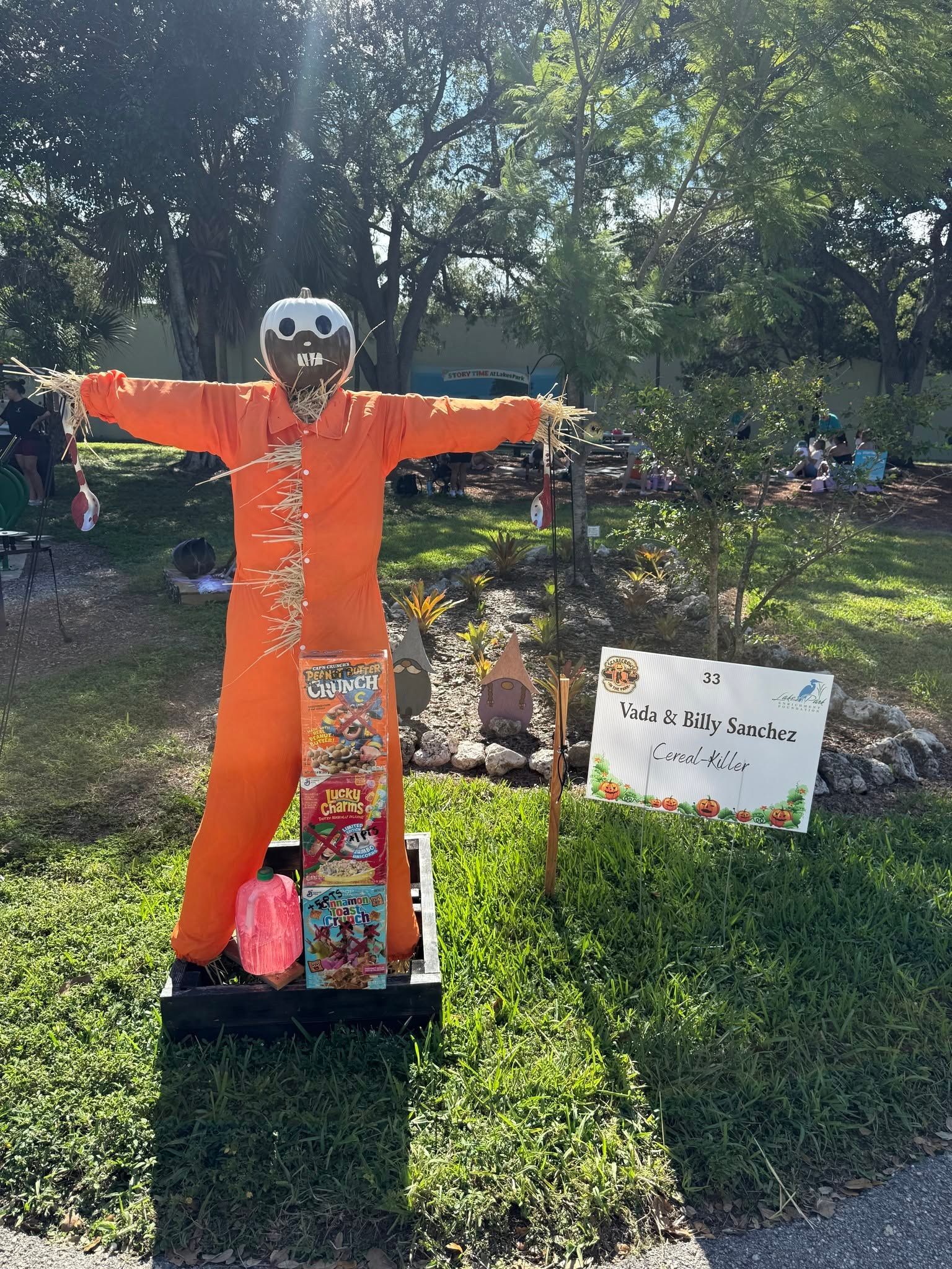 Orange scarecrow with arms outstretched in a grassy park, with trees and signs in the background