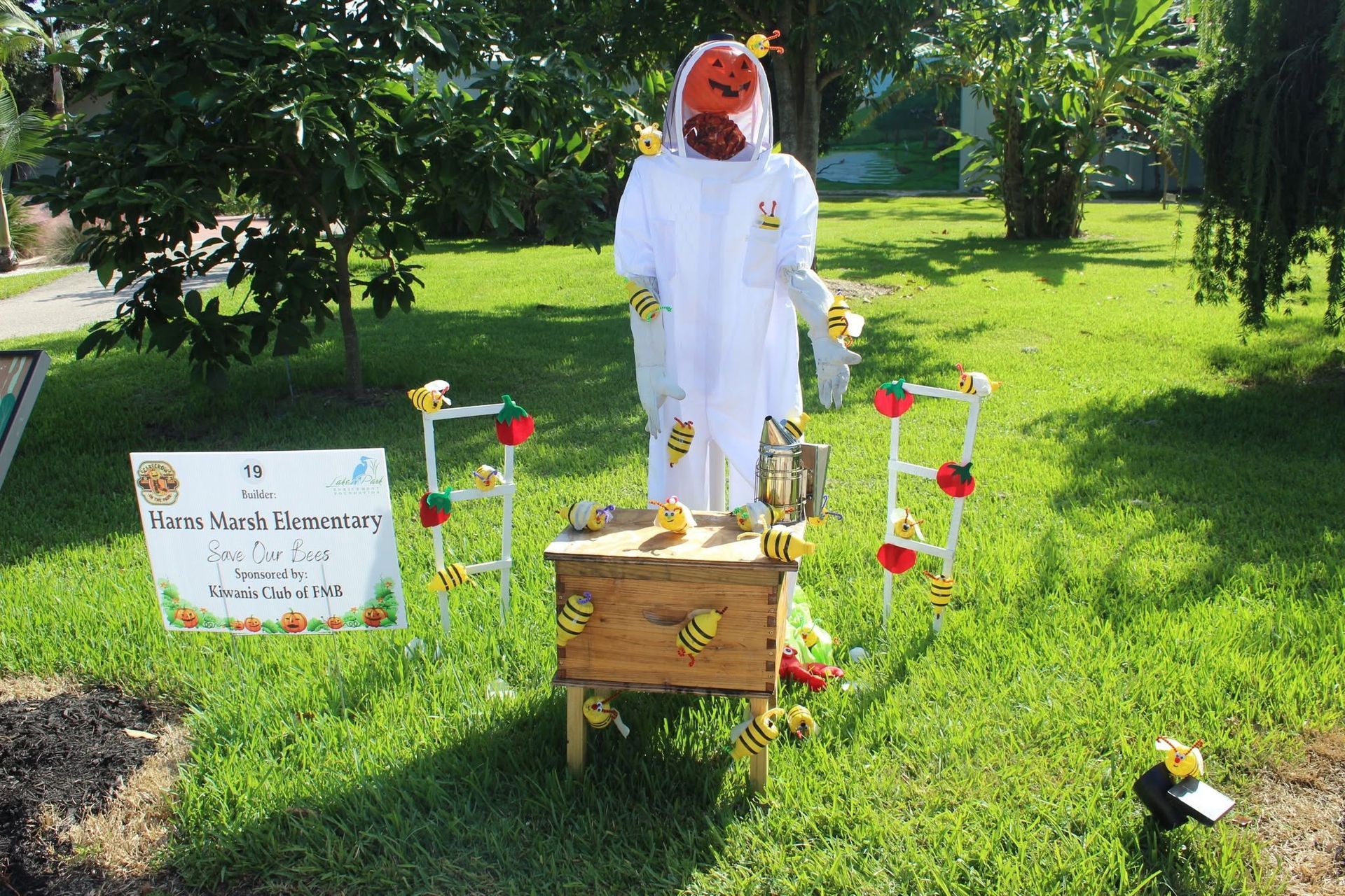 Scarecrow in a white outfit stands in a sunny garden display with flowers and a small sign.