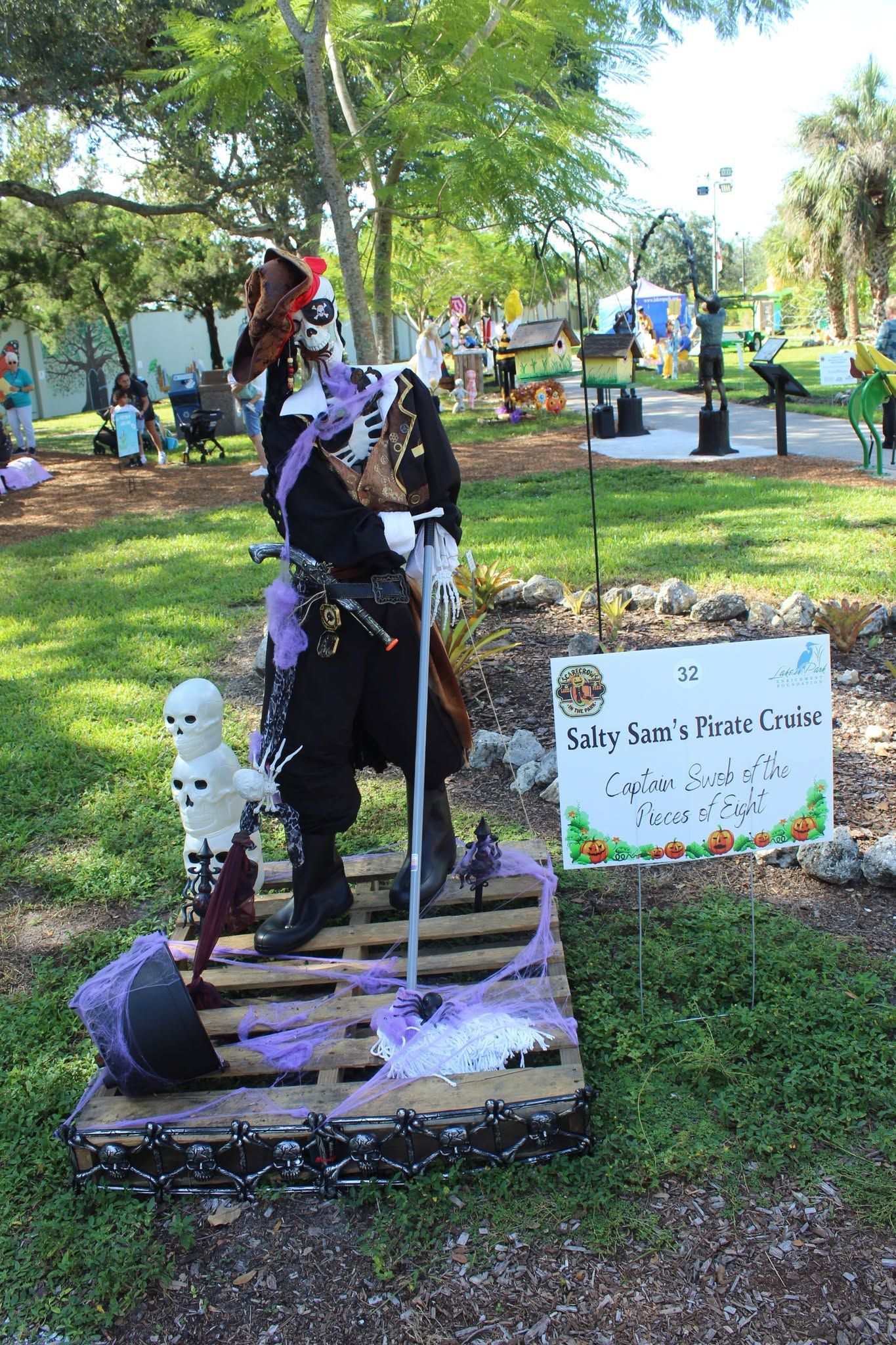 Outdoor Halloween graveyard display with skeletons, a witch figure, and a spooky sign in a park