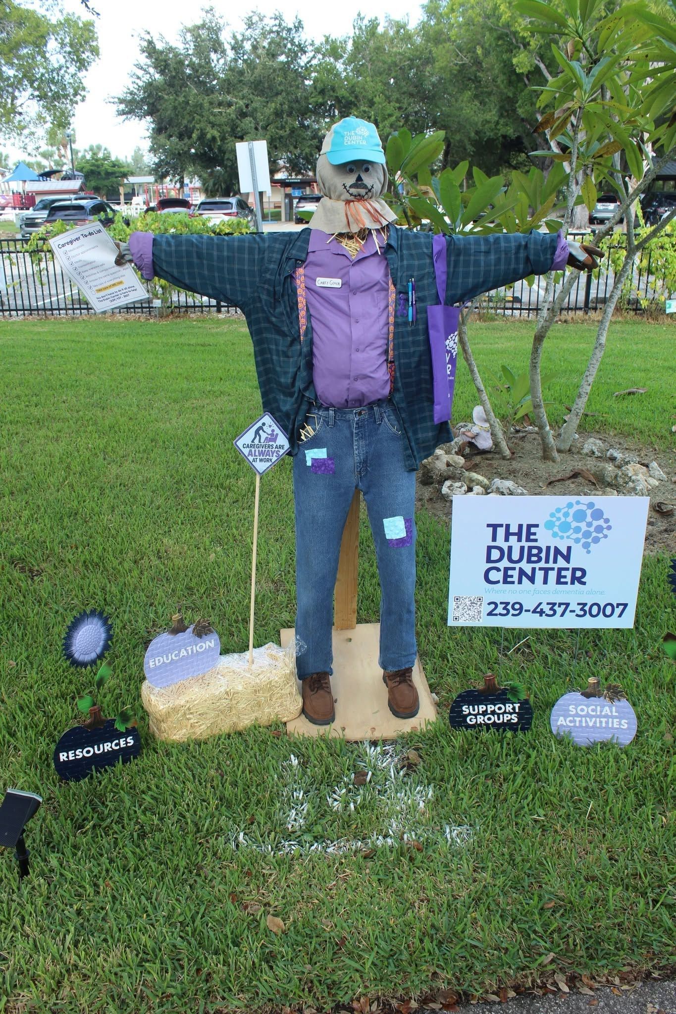 Life-size scarecrow cutout in a park, wearing a hat and dark jacket beside a sign for “The Dent Center.”