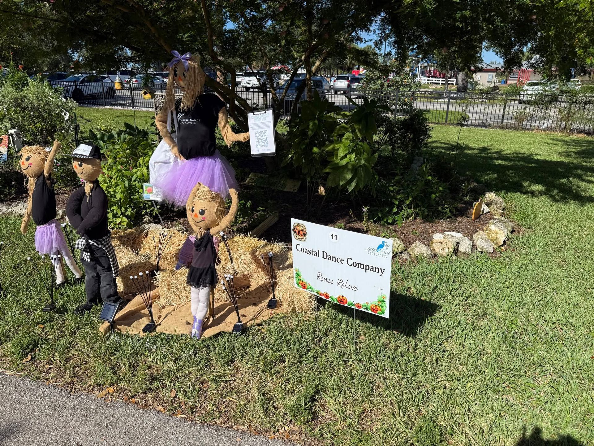 Halloween scarecrow display on a grassy roadside, with a sign and stuffed figures under a tree.