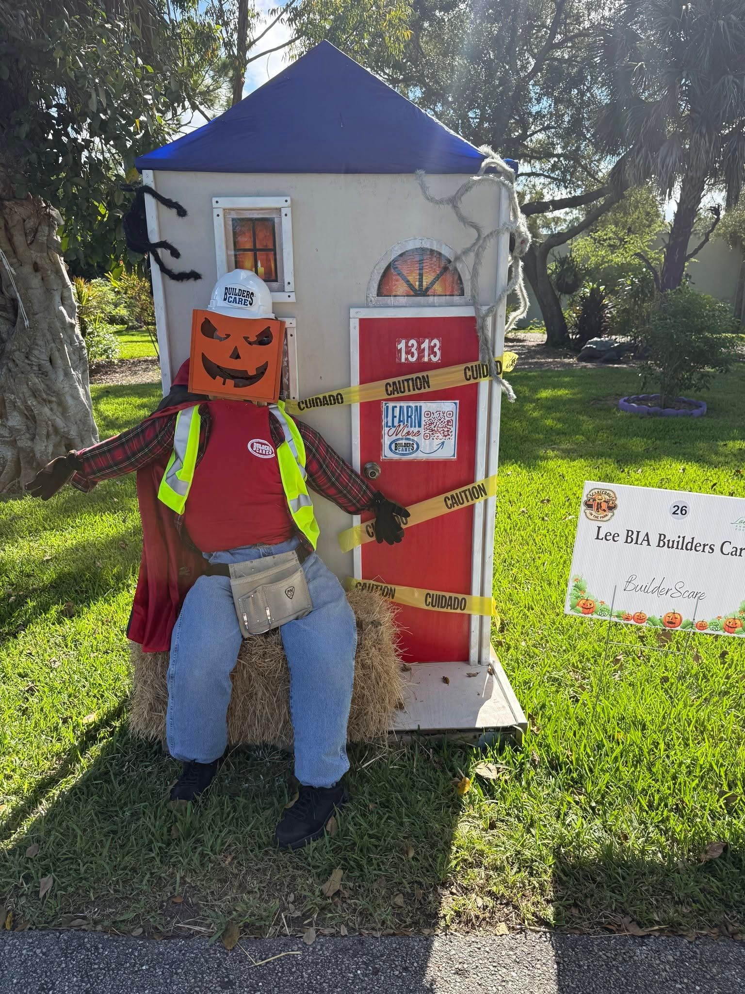 Person in Halloween costume standing by a small decorated house prop on a sunny lawn.