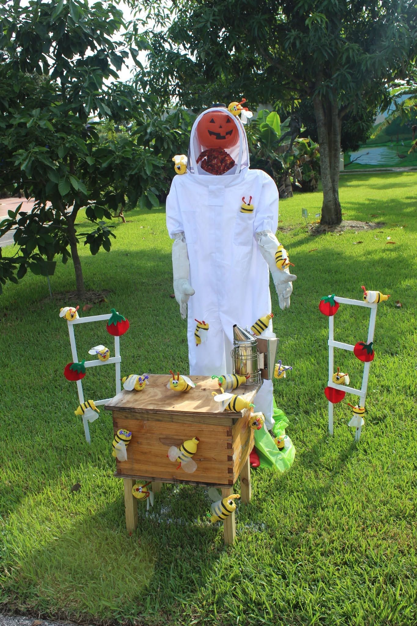 Scarecrow in a white coat stands in a grassy garden beside a decorated wooden hive-like box.