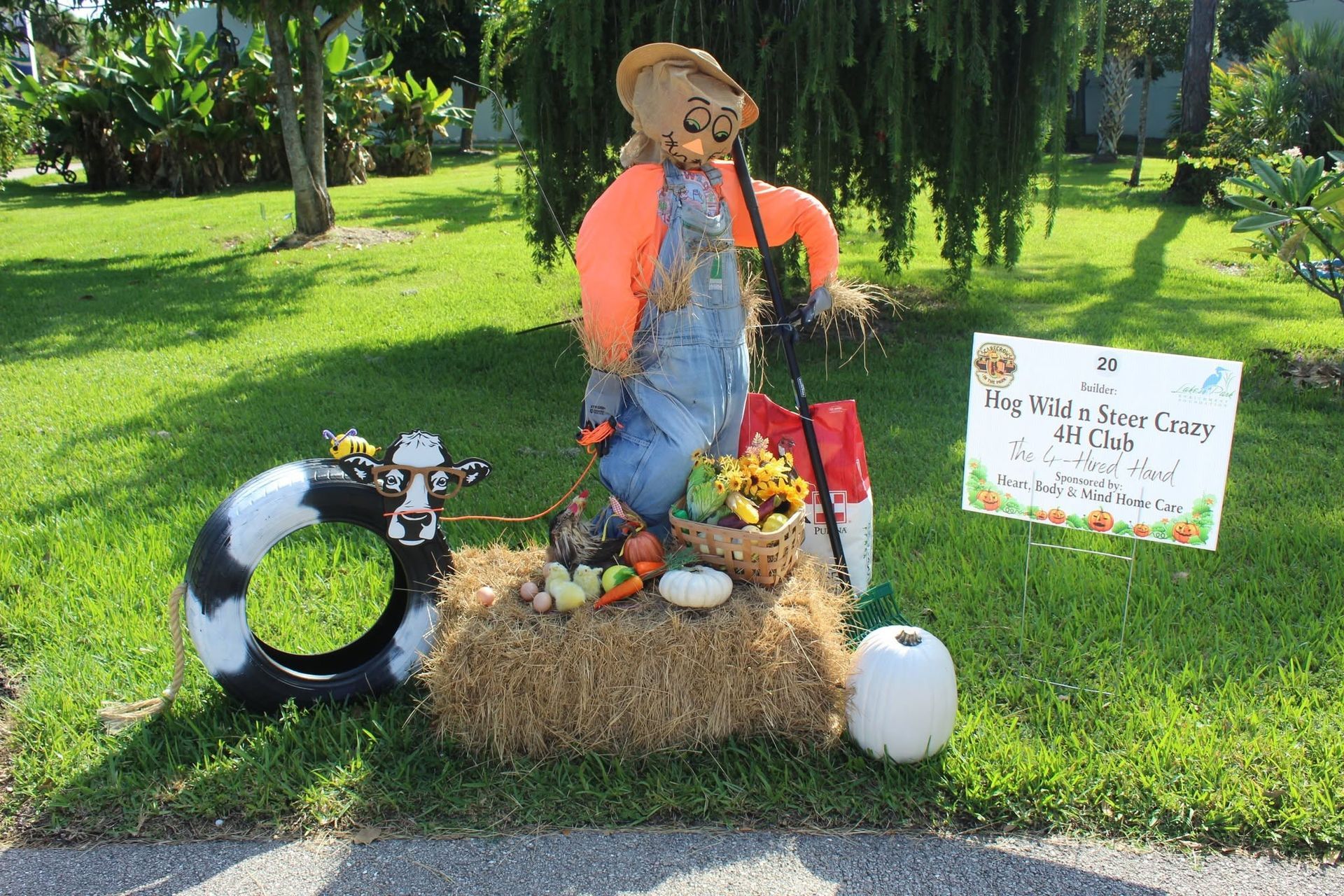 Scarecrow display in a grassy garden with hay bale, pumpkins, flowers, and a sign.