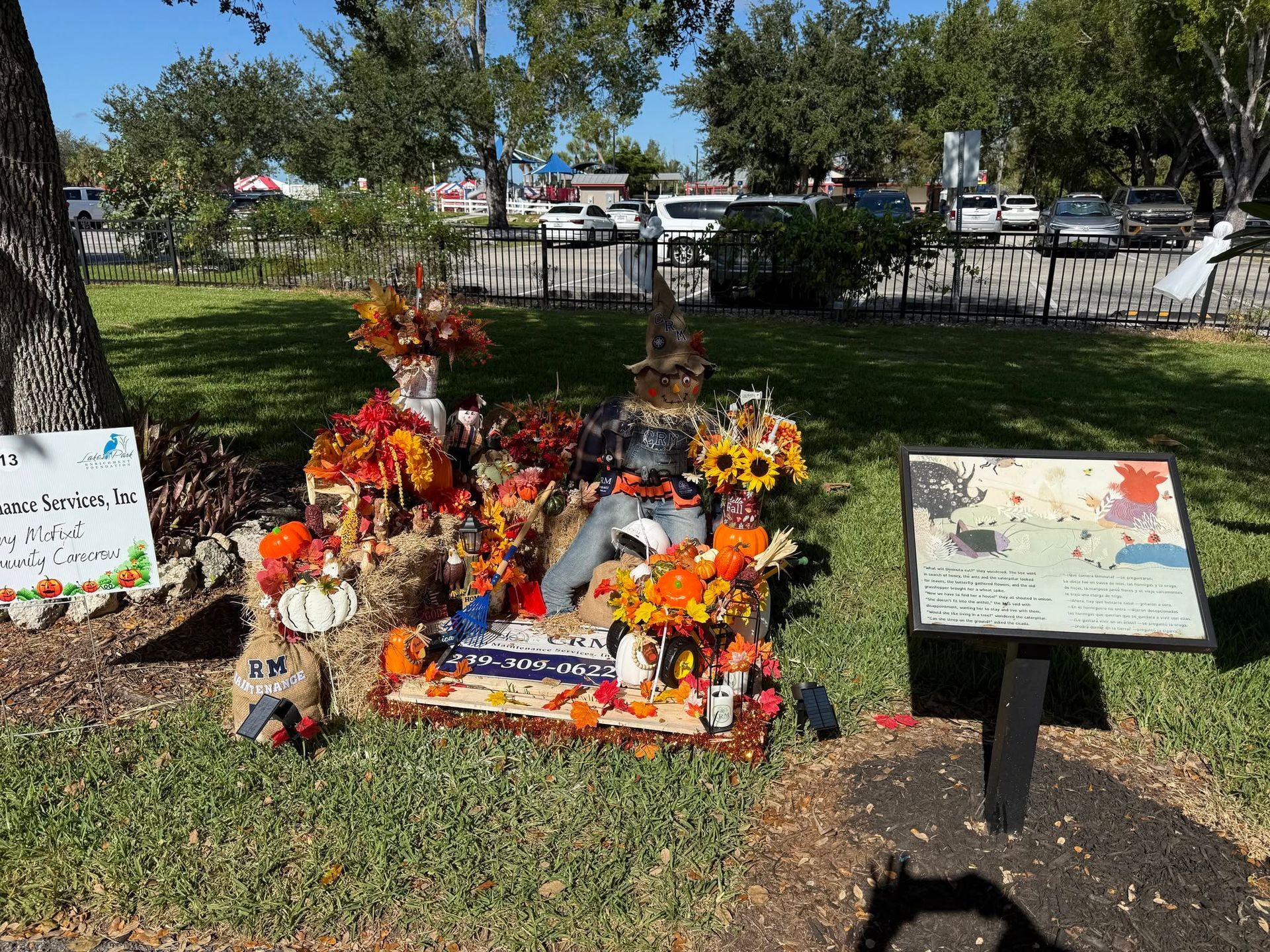 Memorial display with flowers and stuffed animals in a park, beside a sign and informational plaque
