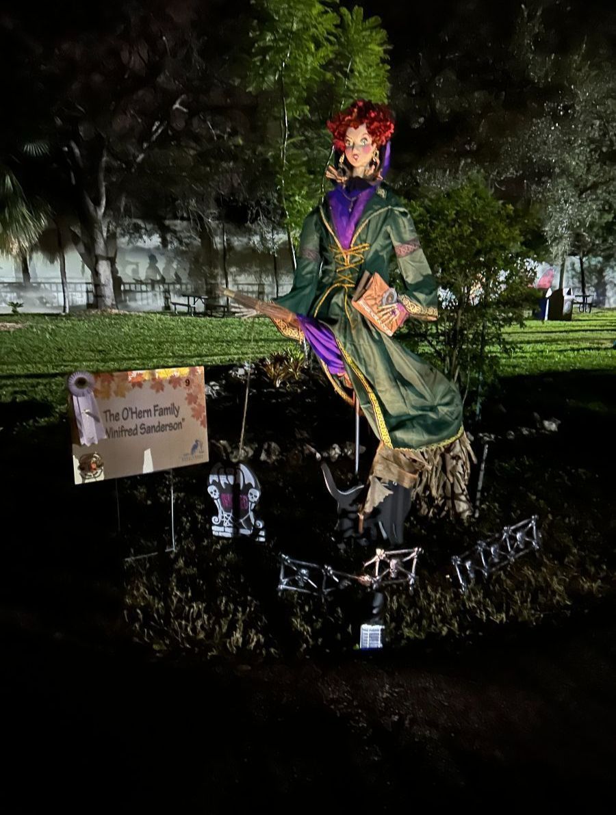 A Halloween display featuring a witch in a green dress holding a book and posing in a yard with a sign.