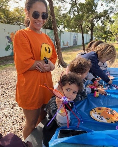 A woman in an orange shirt is standing next to a group of children playing with toys.