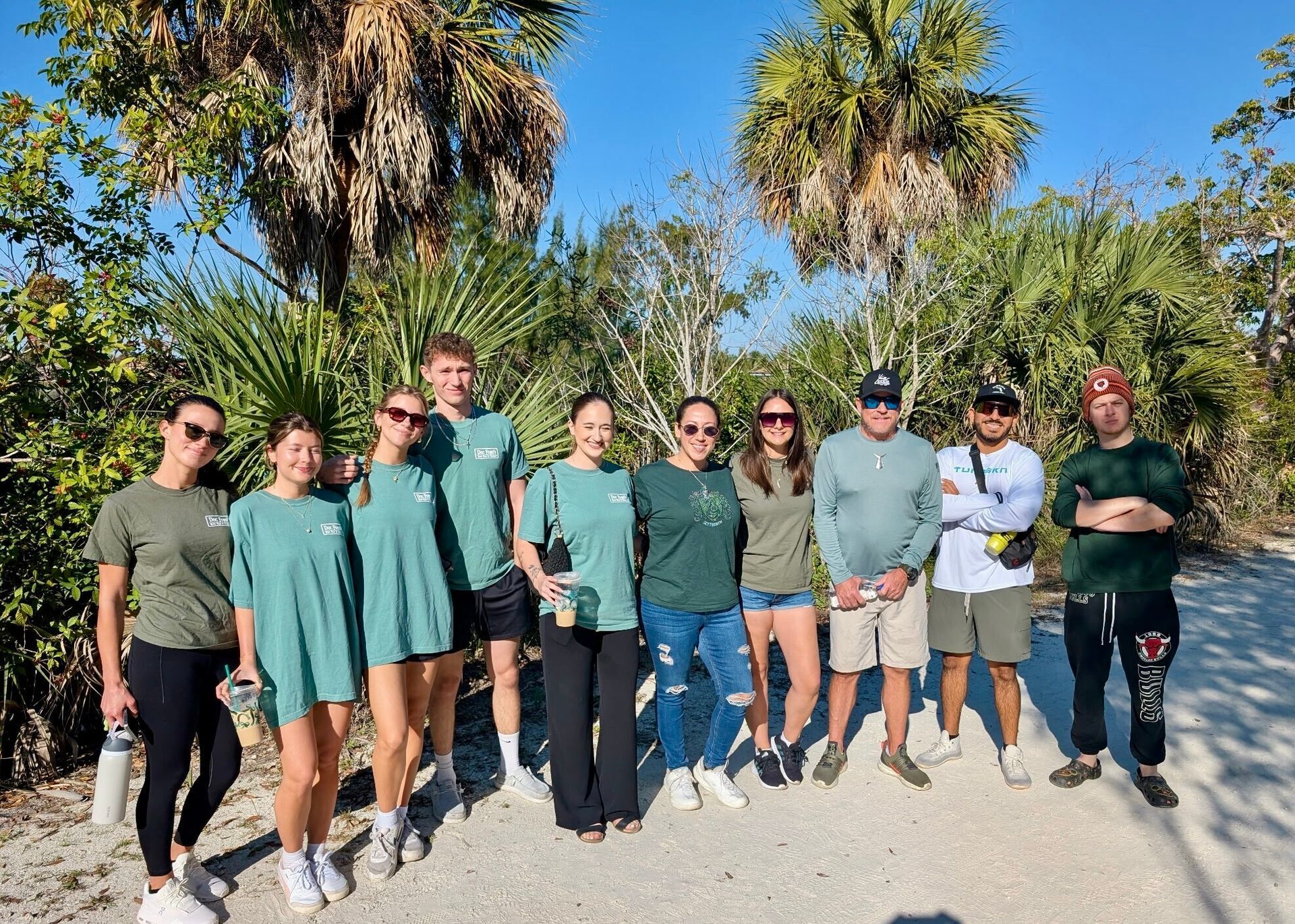 Group of people smiling, standing on a sandy path with trees, wearing casual clothes. Sunny day.
