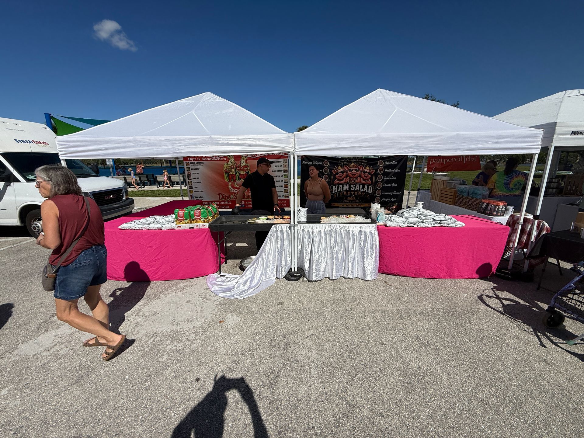 Outdoor market booth under white tents with bright pink tablecloths and a white drape, with a person walking by