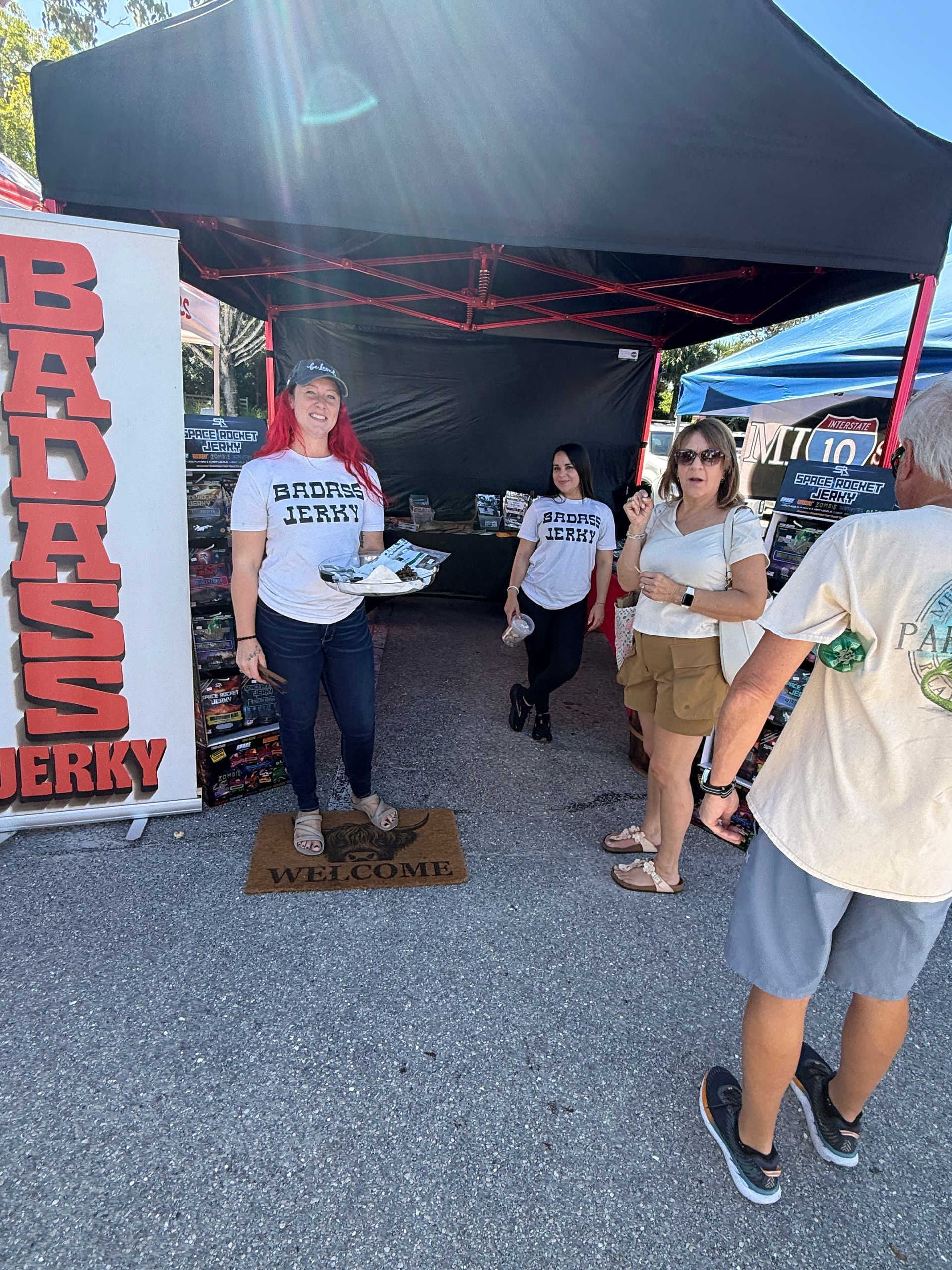 People at a jerky booth under a black canopy, with a red sign reading “Badass Jerky.”