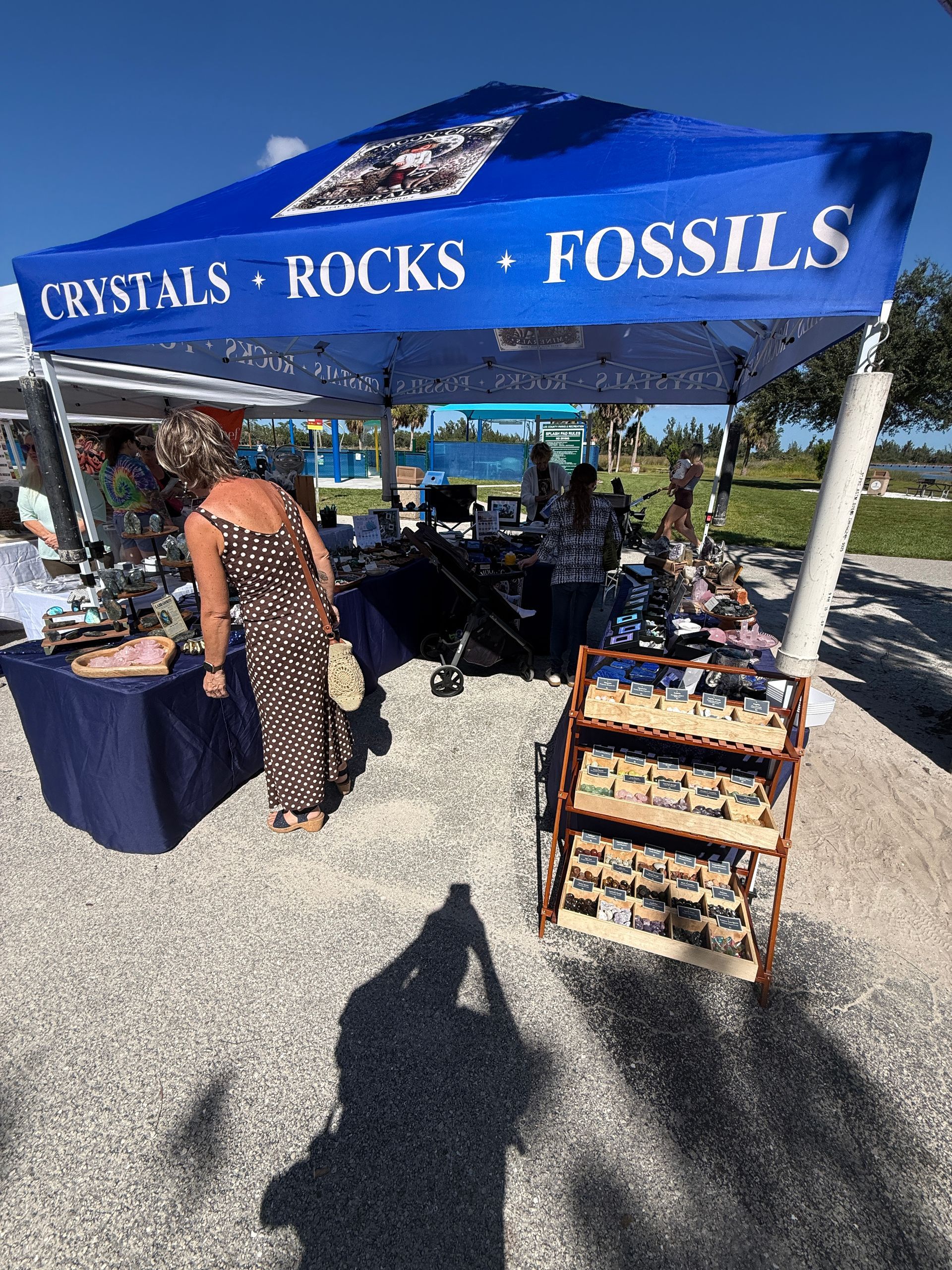 Blue “CRYSTALS • ROCKS • FOSSILS” market tent with displays and a shopper outdoors in bright sun