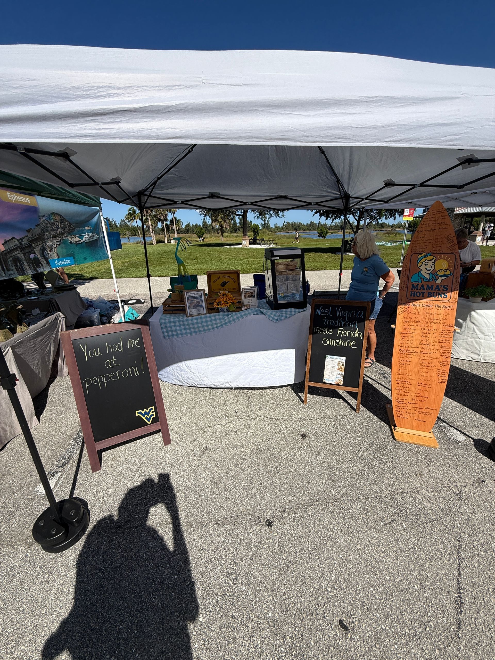 Outdoor market booth under a white canopy with tables, chalkboard signs, and a vendor in the background.