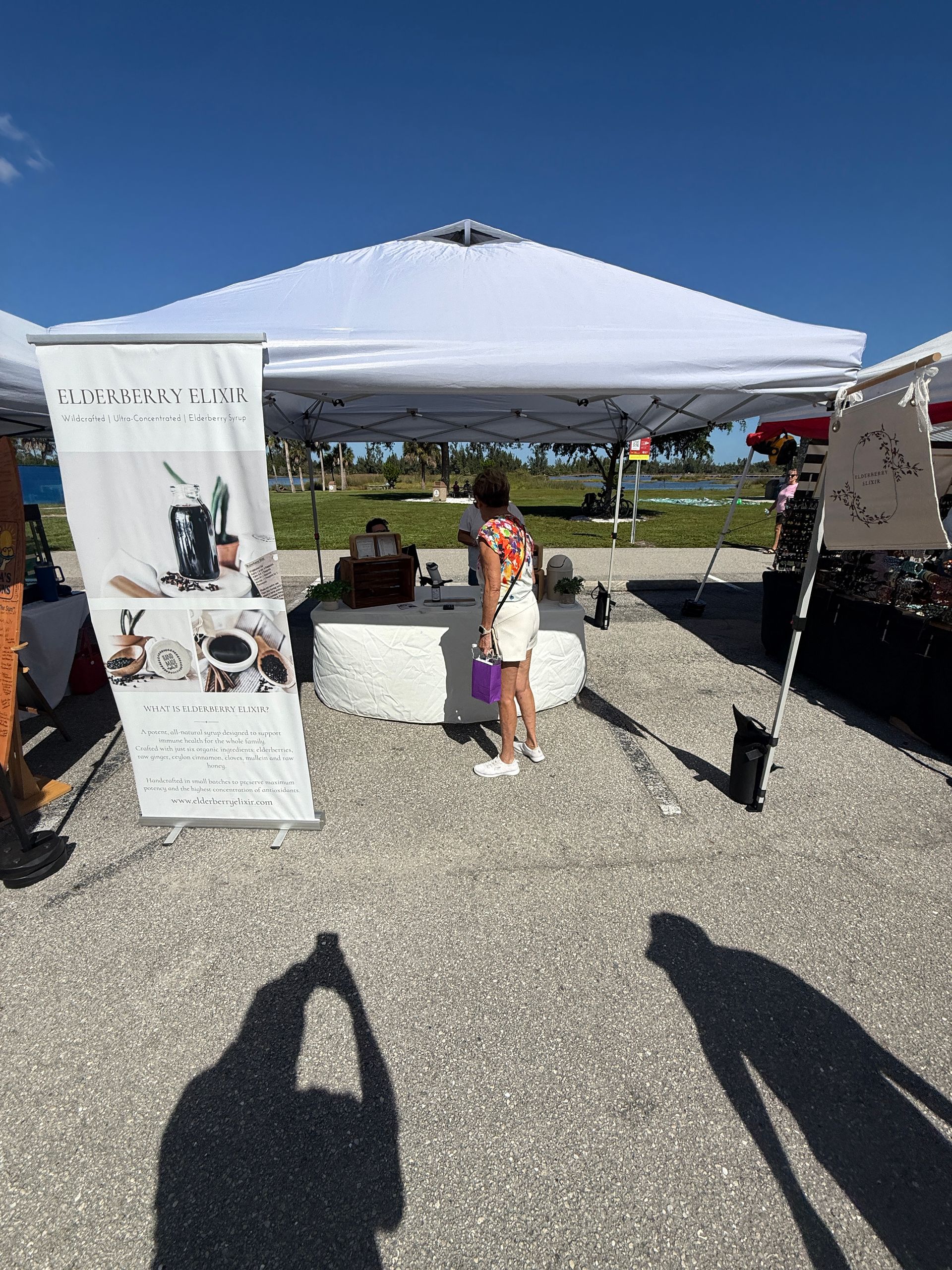 Outdoor market booth under a white canopy with displays, a child in front, and a sunny hillside view behind.