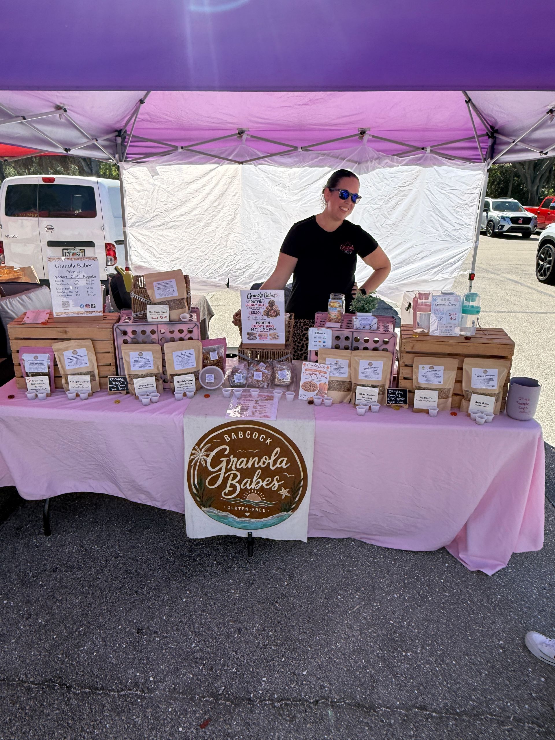 Market stall with pink canopy and packaged baked goods on display, vendor standing behind table