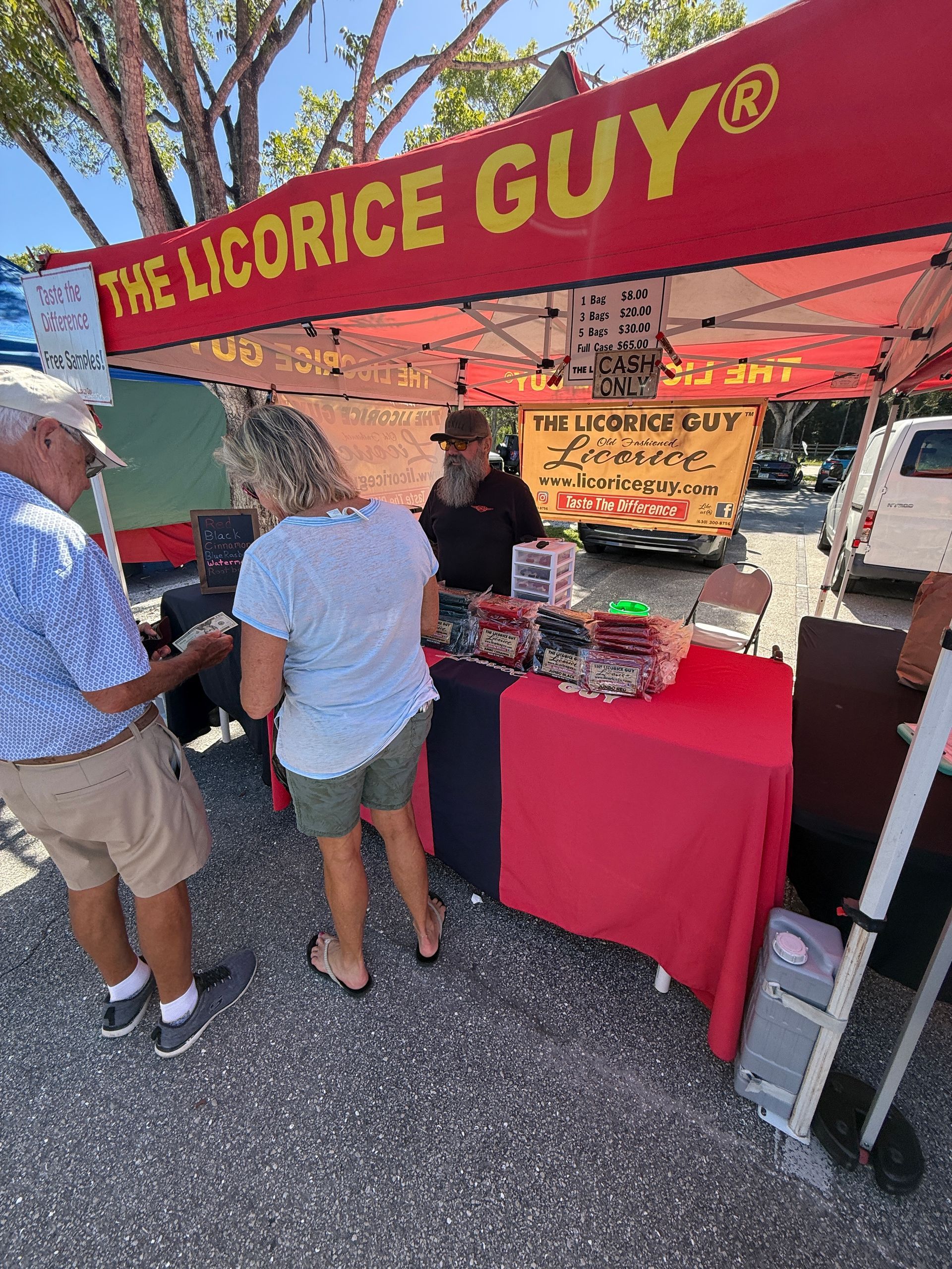 The Licorice Guy market stall with red canopy and two customers browsing candy displays