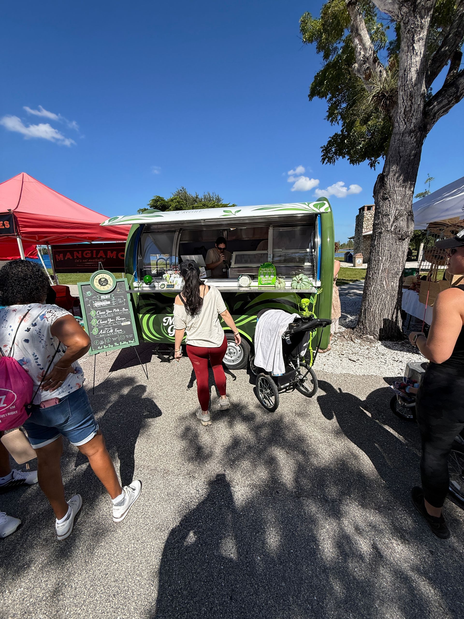 Outdoor market with a green food truck, red canopy, and people walking under a sunny blue sky