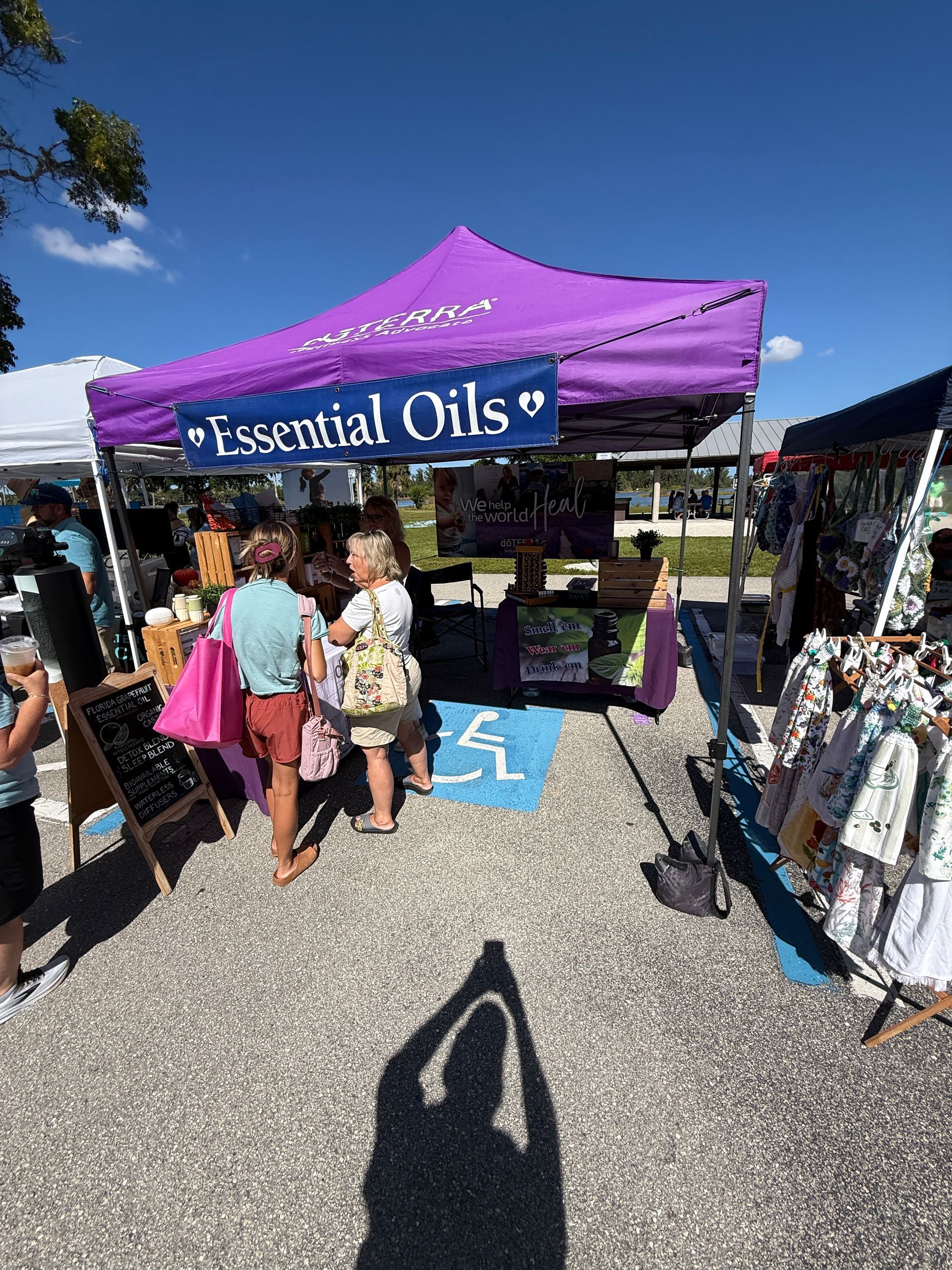 Purple “Essential Oils” market tent with shoppers browsing outdoors on a sunny day