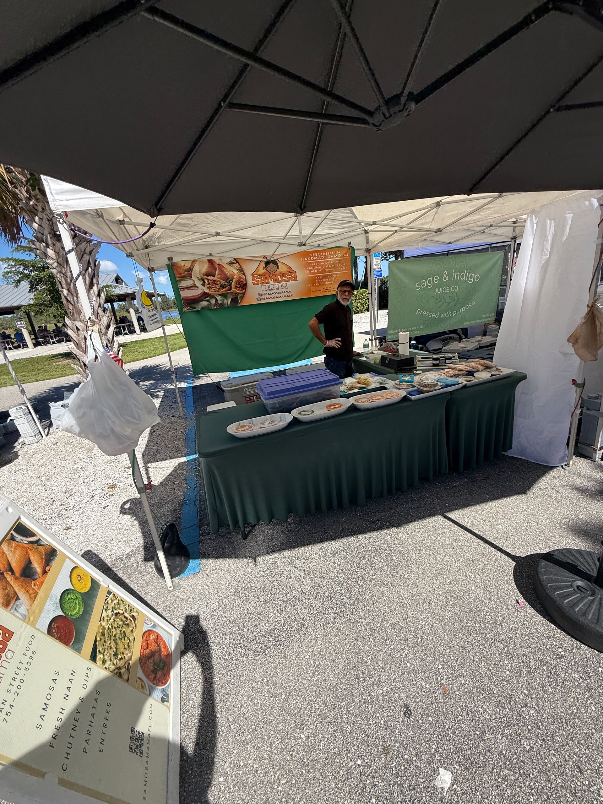 Outdoor market stall under a canopy, with green tablecloths, crafts, and a vendor behind the table.