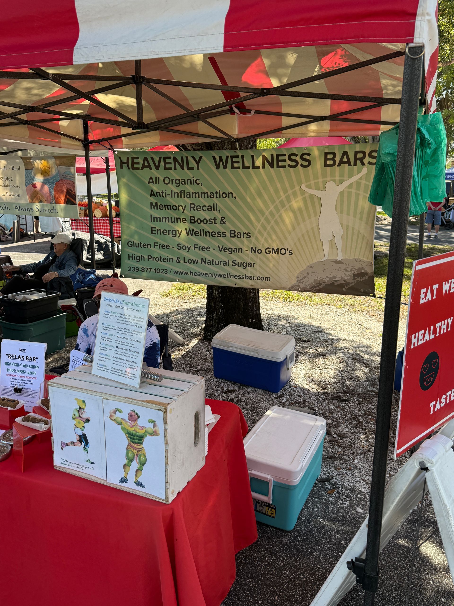 Market stall for Heavenly Wellness Bars under a red canopy, with signs and product boxes on a table.
