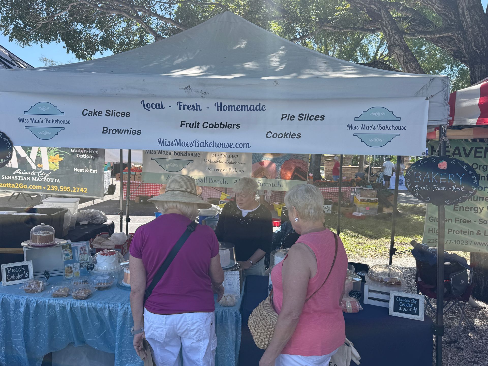 Farmers market booth under a white tent with craft items and two customers browsing.