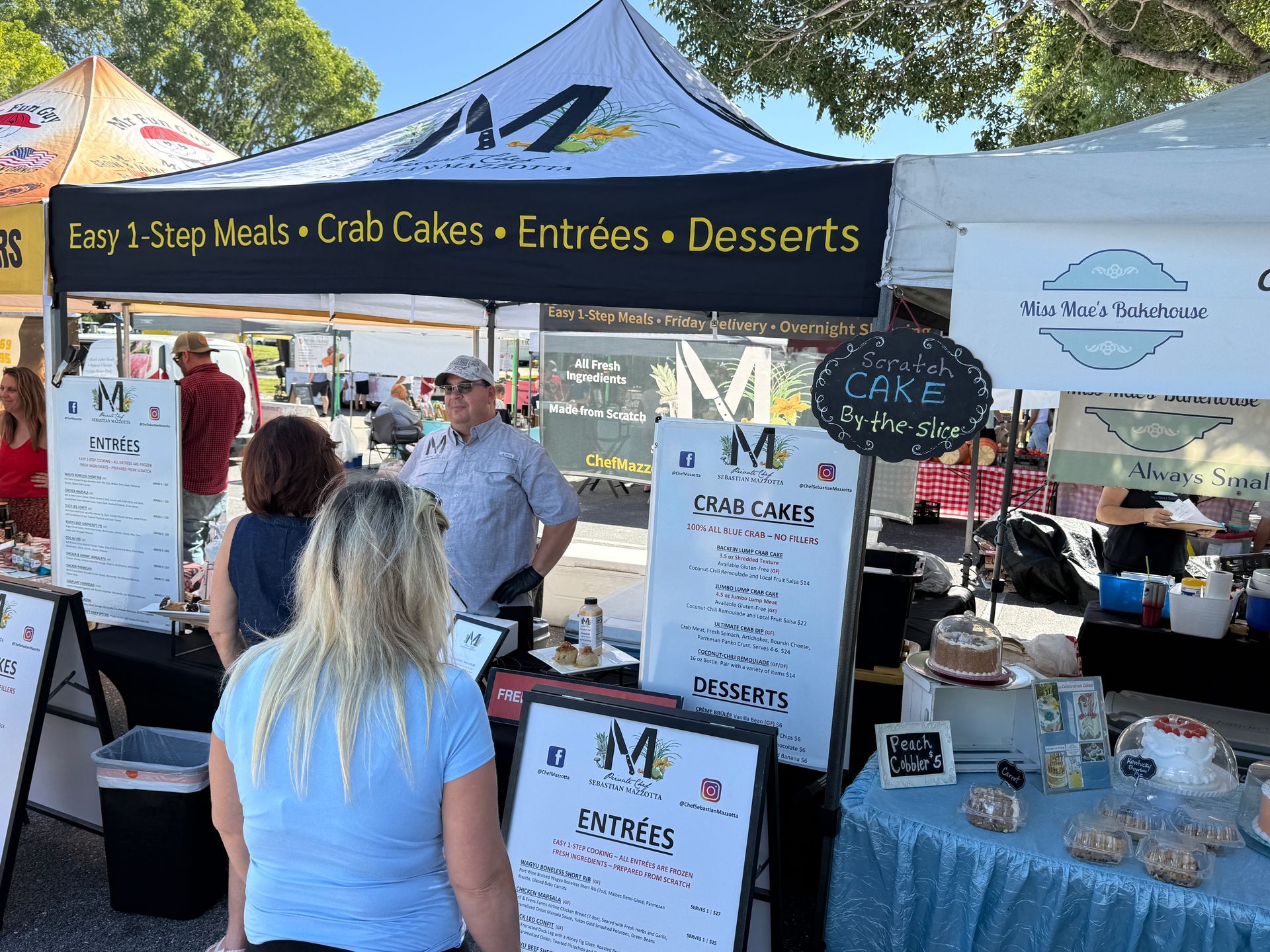 Outdoor food stall with black awning offering crab cakes, entrées, and desserts, with a menu sign and customers nearby