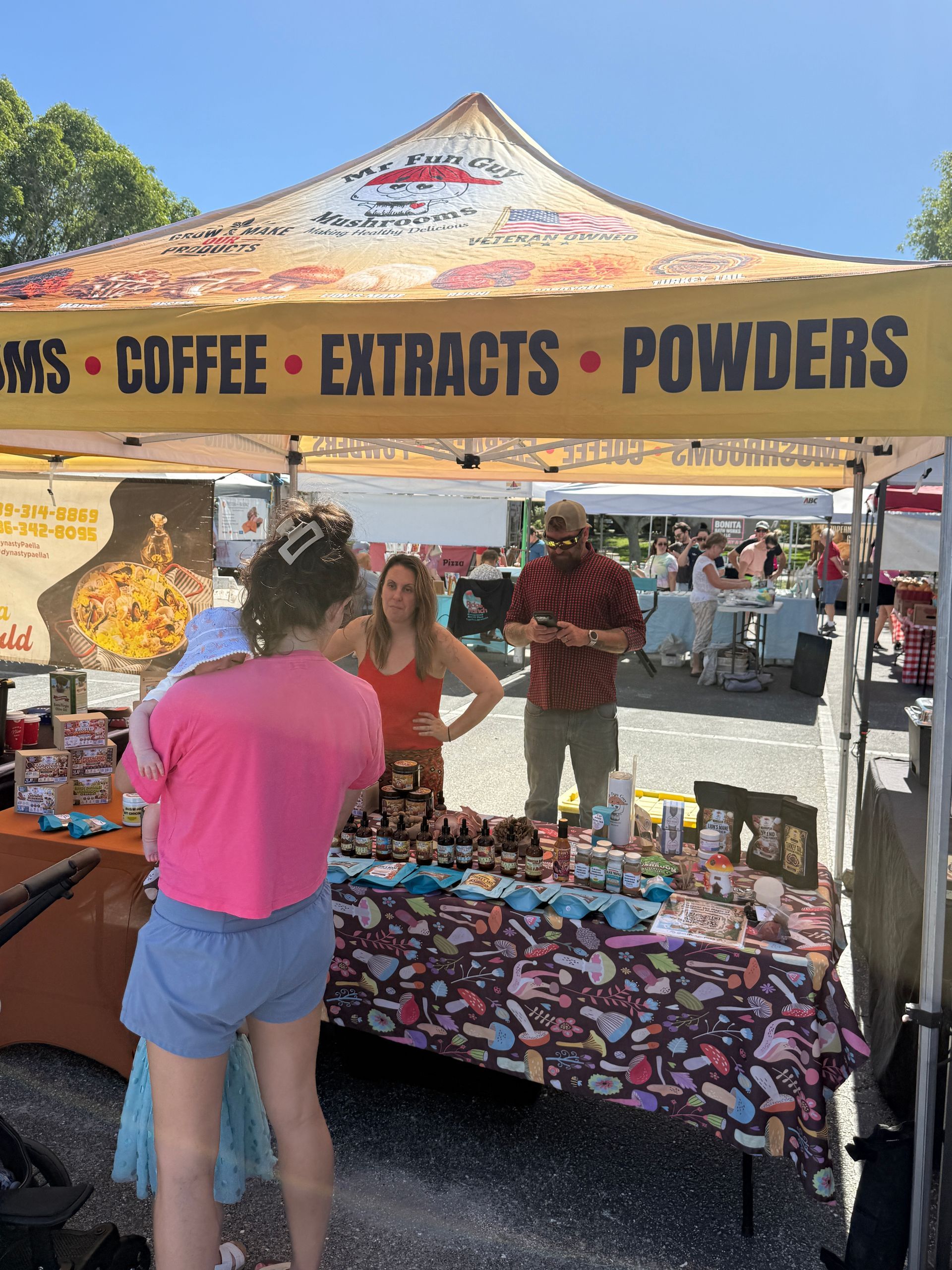 Market stall under a canopy selling coffee extracts and powders, with shoppers browsing colorful items
