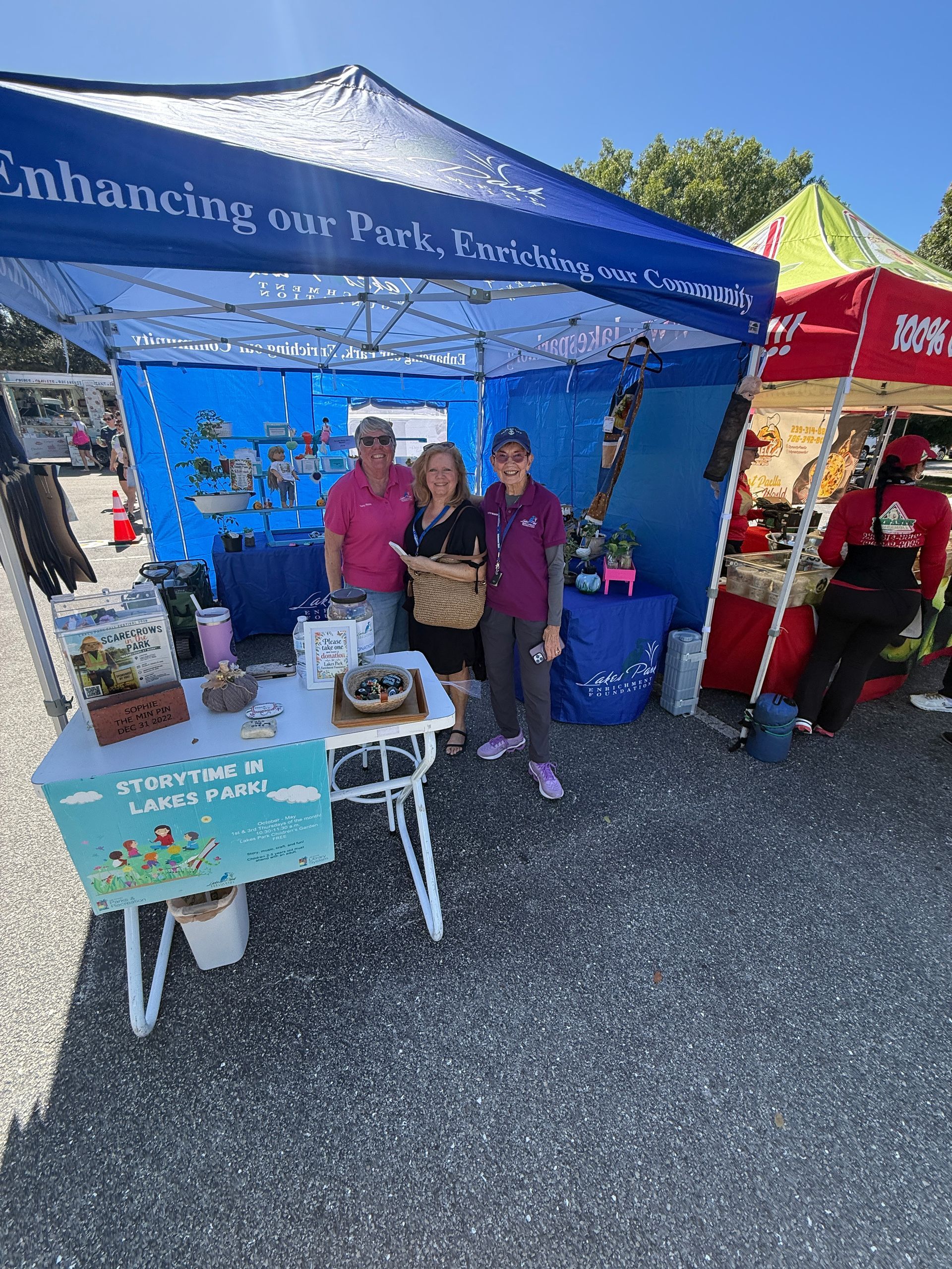 People at a blue outdoor market booth with potted plants and a teal sign under a canopy