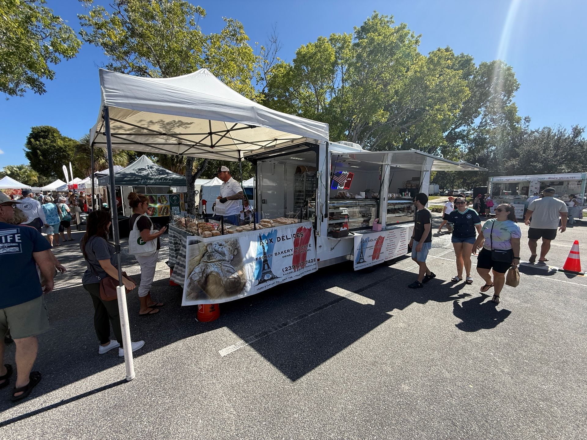 Outdoor market food stall with people standing nearby under a white canopy on a sunny day